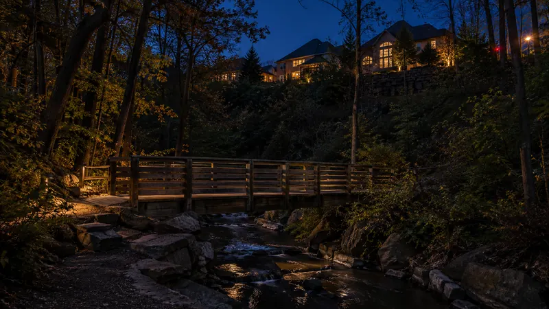 A wooden footbridge over a creek on a forested trail with newer homes above the slope in West Oak Trails at dusk
