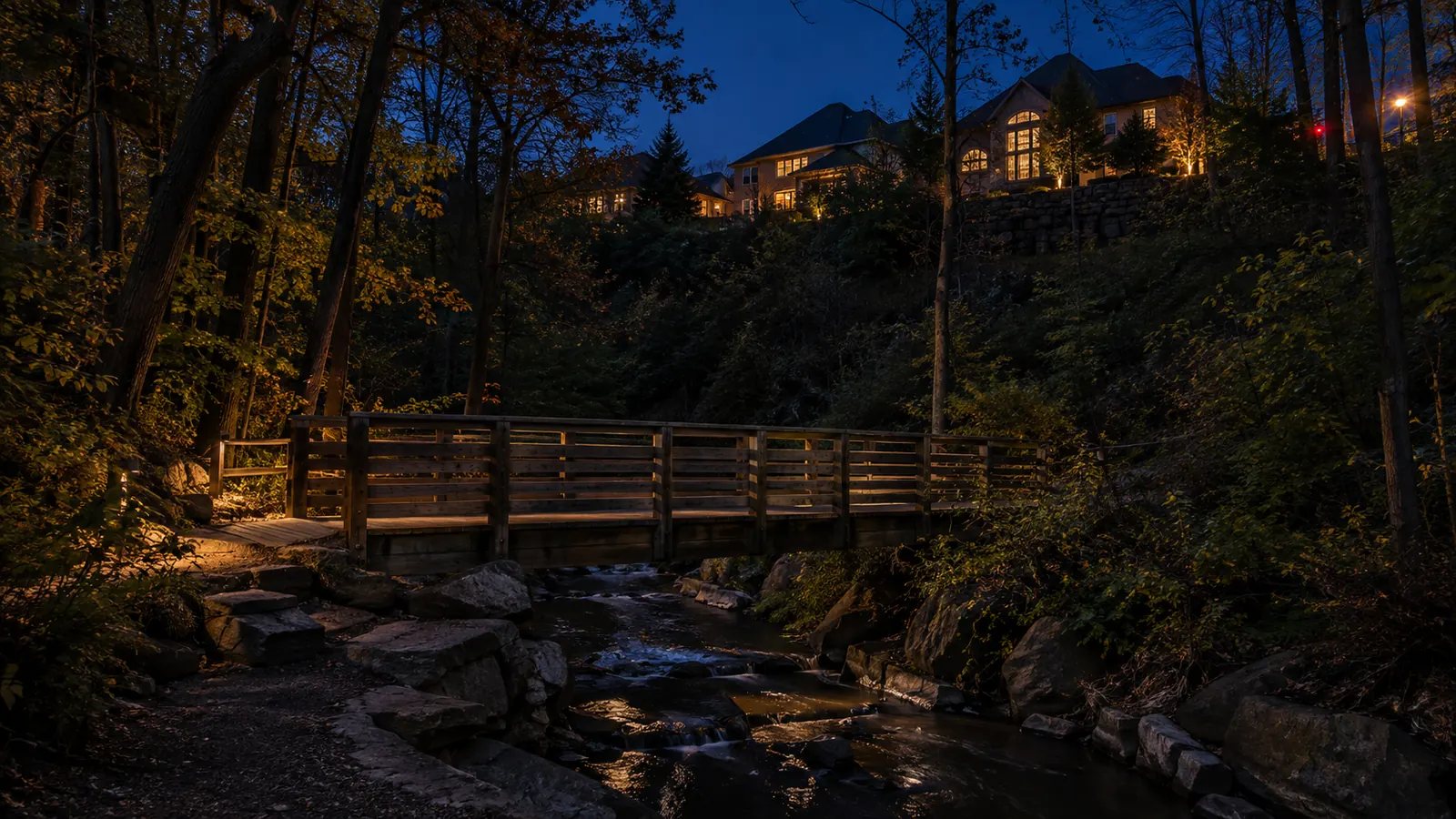 A wooden footbridge over a creek on a forested trail with newer homes above the slope in West Oak Trails at dusk